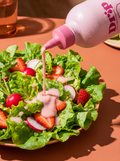 A close-up of a salad bowl with green lettuce, sliced strawberries, radishes, and a pink dressing being poured from a pink squeeze bottle labeled Drip on a warm peach table.