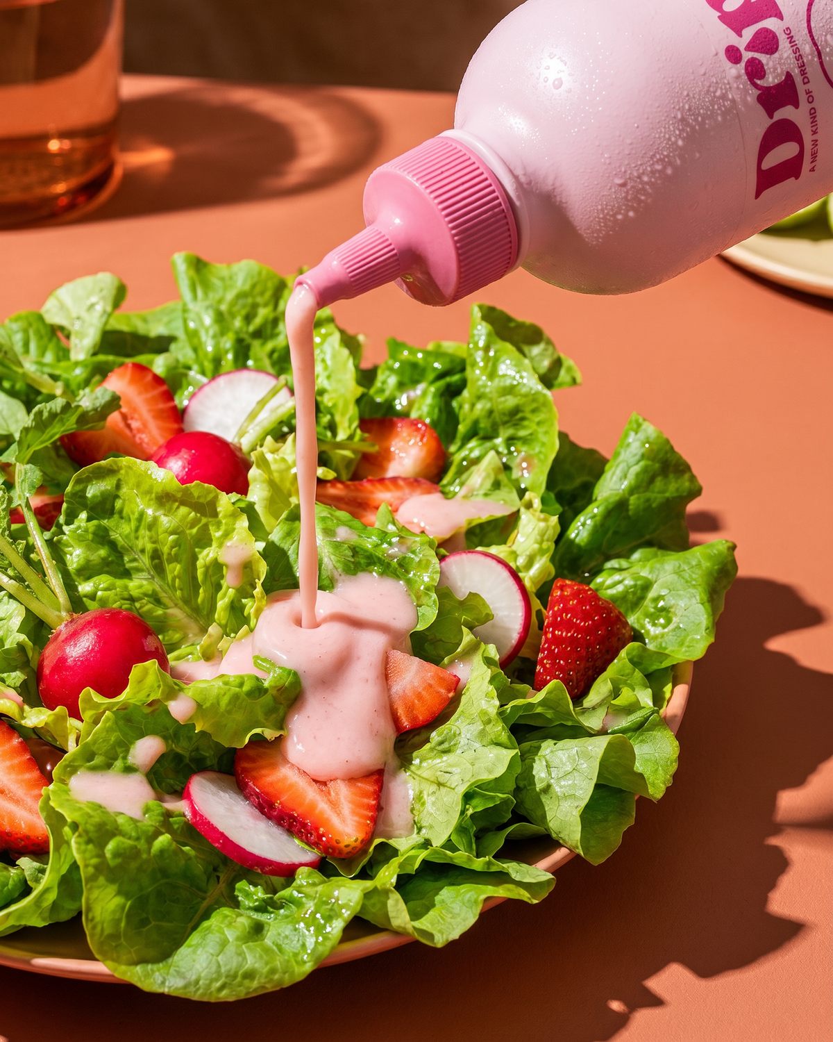 A close-up of a salad bowl with green lettuce, sliced strawberries, radishes, and a pink dressing being poured from a pink squeeze bottle labeled Drip on a warm peach table.