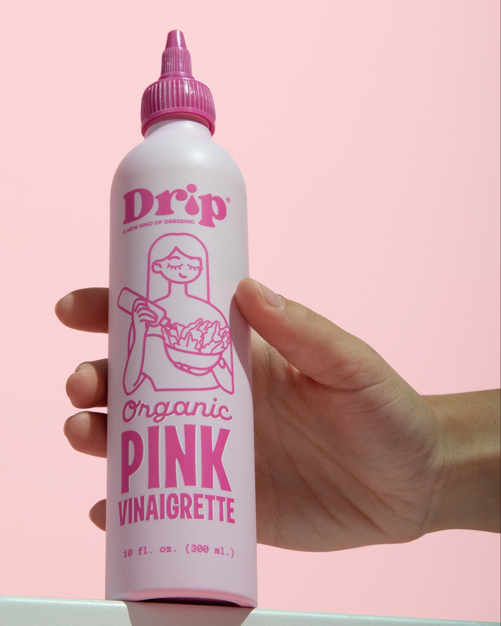 A hand holding a white squeeze bottle of Drip Organic Pink Vinaigrette against a solid pink background.